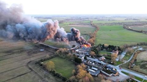 Aerial view of the fire showing flames and a large column of dark smoke drifting with buildings in the middle surrounded by green fields.