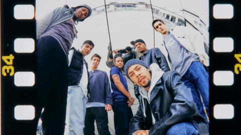 A group of men are standing, with one kneeling, all looking down at a camera with a fish-eye lens. The picture is on a roll of film.