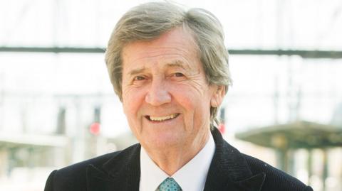 Melvyn Bragg smiling and posing in a suit