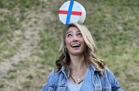 A woman with blonde hair and wearing a blue denim jacket smiles as she balances a round double gloucester cheese on her head