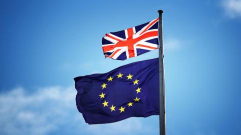 A small red, white and blue British Union Jack blows in the breeze above a a European Union flag, which consists of a ring of yellow stars against a block blue background. The sky in the background is clear.