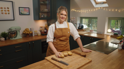 A woman wearing a white shirt and brown apron stands in a kitchen. In front of her on the bench is a chopping board with three potatoes on it