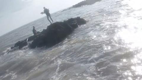 The silhouette of two children on a rock in the middle of a choppy sea. One is sitting down while the other stands. They are clearly stranded.