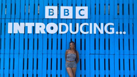 Tianna standing under a sign saying ý Introducing with a completely blue backdrop