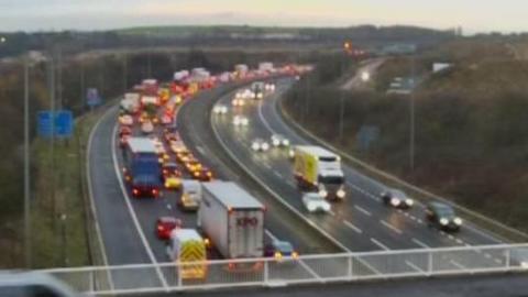 A view of the motorway shows long queues on the westbound carriageway. Traffic is flowing on the opposite side.