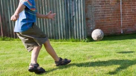 A child is kicking a ball towards a brick wall where another child is waiting to catch it. He is wearing sandals, shorts and a T-shirt. This is a stock picture.