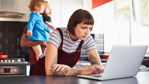 A mum reading leaning down on a table looking at a laptop reading the Bitesize home education newsletter while dad holds up child in the kitchen background.