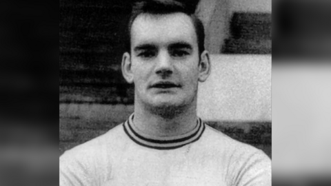 A black and white photo of Ken Houghton looking at the camera. He has short dark hair and is wearing a white t-shirt with a striped neckline. 