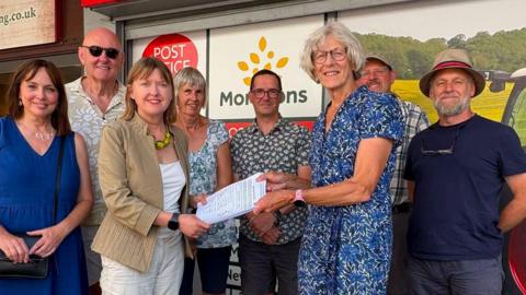 MP Sarah Gibson and councillor Celia Stevens smiling as they stand outside the Post Office branch holding a piece of paper which appears to be the petition. They are standing with several other people who are smiling at the camera.