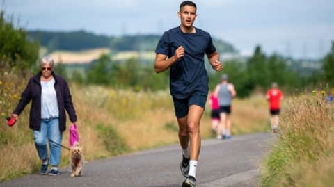 An image of Amol Randhawa, who is running the length of England in 13 days in honour of his mother who died. Amol is wearing a navy t-shirt and shorts, and can be seen running on a path in a park.