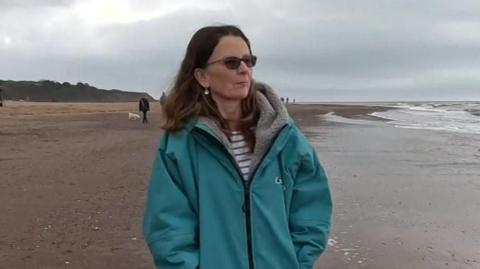 A woman wearing a long blue jacket walking along the beach with the sea to her right.