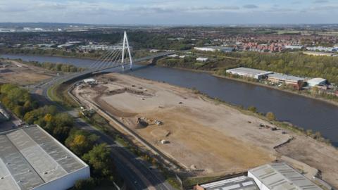 An aerial photo of the Crown Works site which is next to the River Wear. The land on the left of the river has been flattened and is a sand colour. There are a number of vehicles on the land towards the back. The white Northern Spire bridge goes across the river beyond the Crown Works site. On the other side of the river there are warehouses and houses in the distance. The sky above is cloudy.