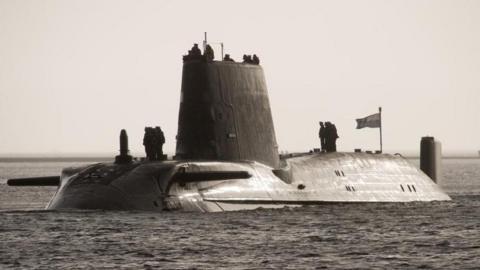 A file photo from 2009 shows HMS Astute in a body of water - troops stand on top of the vessel and a flag flies in the wind.