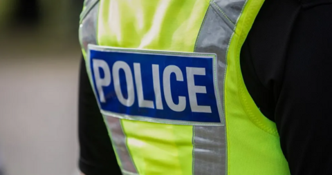 A close up picture shows a police officer's back. The officer is wearing a black top and a high-vis vest with the word "police" on the back. 