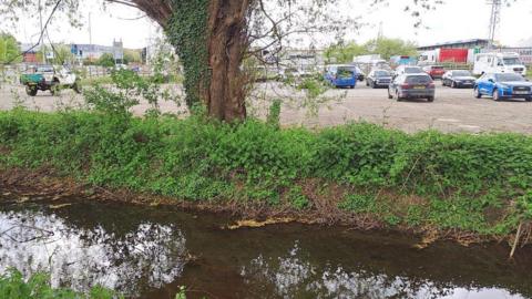 A shallow brook runs adjacent to a small car park with Hereford FC's stadium in the background. 