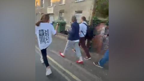 A woman holds her phone while videoing a man and a woman walking along the street, in a blurry screenshot from mobile phone footage.