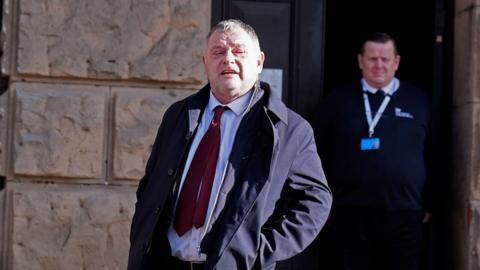 MP Mike Amesbury leaving court in Chester. He has a five o'clock shadow, he is wearing a dark blue jacket, over a white shirt and red tie.