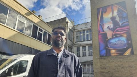 Artist Mohammed Ali Aerosol is pictured outside a building bearing a mural which shows a car with the wing mirror projecting an image of a building