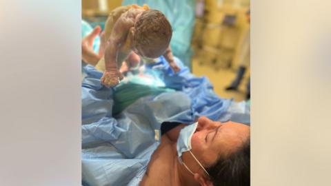 Dr Leila El Alti, a woman with dark hair, lying on a hospital bed while wearing a blue surgical gown and blue face mask, holding her newborn baby into the air. 