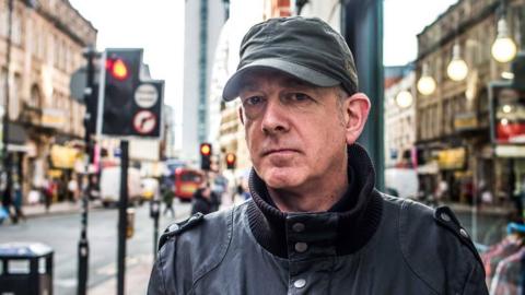 Dave Haslam, a man wearing a black coat and a dark-coloured baseball cap, is stood near some traffic lights in a city centre.
