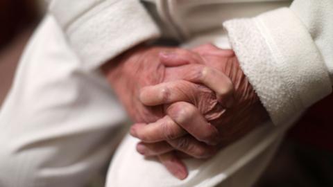 An elderly woman's hands resting on her knee. She's wearing a white fleece and white trousers.