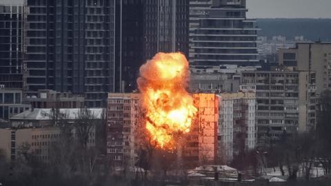 A large explosion and orange fireball rises from an apartment block