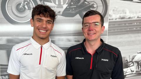 A young man in a white polo shirt and an older man in a black version of the same shirt stand side-by-side with broad smiles.