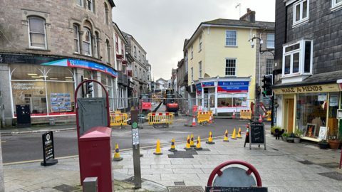 There is a dark grey stone building on the left, on the pavement, which has a shop called Make a Mends. The shop is painted yellow and has a black sign outside. In the centre is a yellow building with another business which has yellow cones and fencing in front of it. The building on the left, another business, also has fencing outside of it with yellow barriers. There is a red box on the left-hand side of the pavement 