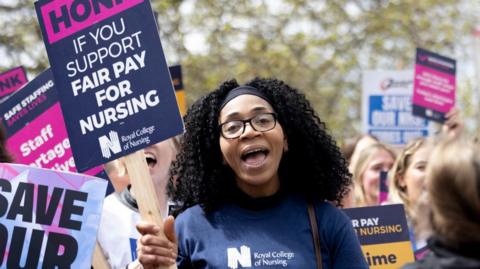 A woman with dark curly hair wearing glasses and a Royal College of Nursing top, holding a sign saying "honk if you support fair pay for nursing" 