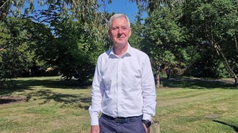 Tim Oliver looking at the camera. He is wearing a grey shirt and blue trousers. Grass and trees in the background.