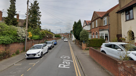 A Google street view screenshot of a residential street of semi-detached houses, with cars parked on one side of the road.