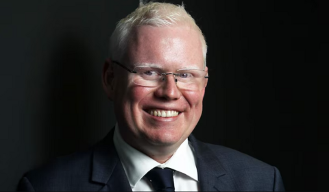 Gareth Ward, smiling at camera, wearing a dark suit and tie and glasses