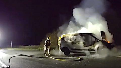 A burning van with a firefighter standing next to it with a hose.