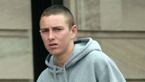 a young man with cropped brown hair and wearing a grey hoodie is standing outside a court building