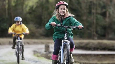 Two children ride on bikes in a rural setting. The child at the front is a girl in a green coat and red helmet, the child behind is blurred and is wearing a yellow coat and blue helmet.