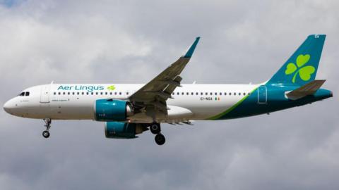 An Aer Lingus plane flying. The plane is white with Aer Lingus branding. The sky is cloudy and grey.