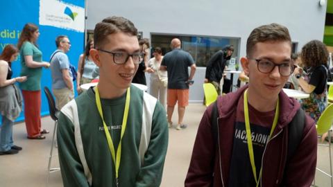 Two twins stand next to each other smiling