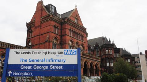 The red-bricked Leeds General Infirmary building with stone dressings and Venetian Gothic windows. A blue and white sign signals the reception area ahead. On the sign, it reads "Leeds Teaching Hospitals" and "The General Infirmary".