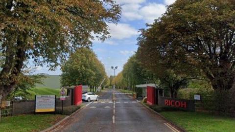 A long road stretches into the distance with green grass and trees on either side of it. There is a brick wall with bright red gates and a red and white barrier for vehicles beyond it. A white car is parked on the left. On the brick wall there is a red sign that reads "Ricoh"