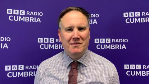 Owain Roberts has short grey/brown hair and is wearing a shirt and maroon tie. He is standing in front of a purple BBC Radio Cumbria branded backdrop.