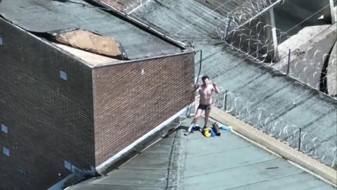 Joe Outlaw, wearing just black boxer shorts, stands on top of a prison roof making peace signs with his fingers. 