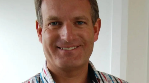 A smiling man in a red, white and blue check shirt.