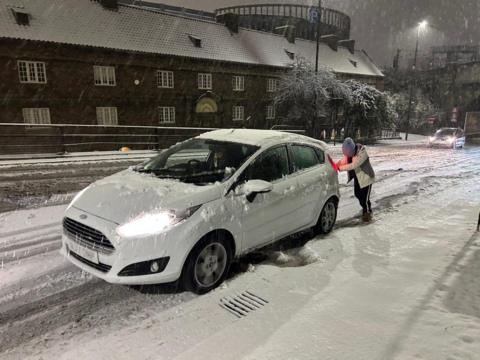 A woman wearing a blue hat and a white jacket pushes a white car along a snow-covered road