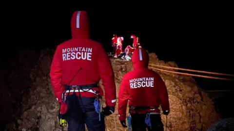 Two members of a mountain rescue team look on at their colleagues on a rocky hill carrying out a rescue operation.