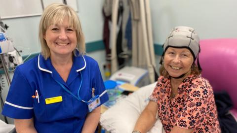 Deputy sister Liz Timms is sitting to the right of patient Sharon Wilkinson in a hospital ward. Liz, who has short blonde hair and a fringe, is wearing a blue uniform with an NHS lanyard. Sharon is wearing an orange floral blouse and a grey chemo cold hat – that looks like a bike helmet – on her head