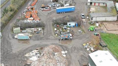 Drone image of a farm yard with a large pile of rubble, and areas of skips, appliances and assorted waste, separated by muddy roads and a number of vehicles
