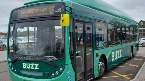 A green bus stopped on yellow hatching in a car park.