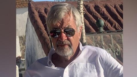 A man with grey hair wearing a white shirt sits at an outdoor table with a glass of white wine. He wears sunglasses along with a bracelet on one wrist.