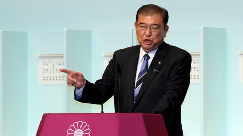 Shigeru Ishiba speaking at a podium, gesturing by pointing his finger, with a blue background