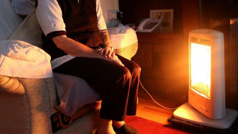 an elderly person sitting in front of a white electric heater- wearing a black and white cardigan and black trousers 